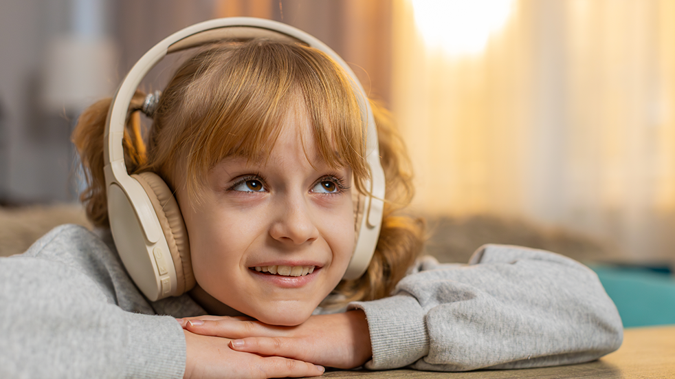 A young girl resting her chin on her hands and wearing big over ear headphones while listening to an audiobook through a smartphone